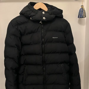 Svart dunjacka från GANT - Säljer en svart dunjacka från GANT i mycket bra skick. Jackan har en dragkedja framtill och en huva med knappar. Perfekt för kalla vinterdagar och håller dig varm och bekväm. Passar både till vardags och lite finare tillfällen. 🖤