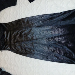 Svart glittrig klänning - Snygg svart klänning med glittrig yta, perfekt för festliga tillfällen. Den har en elegant slits och är tillverkad i ett glansigt material som ger en lyxig känsla. Klänningen är i bra skick och passar perfekt för en utekväll eller nyårsfest.