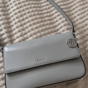 Grå axelväska från DKNY - Säljer en stilren grå axelväska från DKNY i skinn. Väskan har en elegant design med en liten metallbricka och DKNY-logga framtill. 
