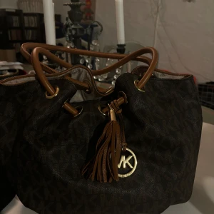 Brun axelväska från Michael Kors - Säljer en snygg brun axelväska från Michael Kors i mycket bra skick. Väskan har ett klassiskt mönster med en dekorativ tofs och guldfärgad logga. Perfekt för både vardag och fest!