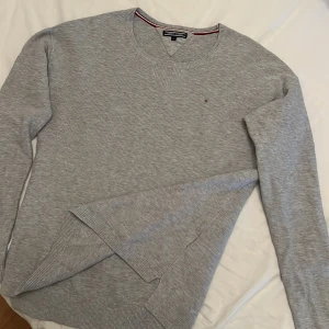 Grå tröja från Tommy Hilfiger - Säljer en stilren grå tröja från Tommy Hilfiger i mycket bra skick. Den är långärmad, ganska tunn och har en liten logga på bröstet. Perfekt för både vardag och lite finare tillfällen. Tröjan är mjuk och bekväm, med ribbade muddar och en klassisk rund halsringning. Stl 176 men liten i storleken så kanske närmare 160. 