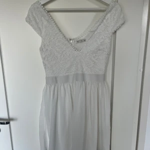 Vit spetsklänning från NLY one  - Elegant vit klänning från NLY ONE med vacker spetsdetalj på överdelen och en mjukt fallande kjol. Klänningen har en v-ringad design och korta ärmar med spets. Perfekt för sommarens fester eller studenten. Dragkedja i ryggen för enkel påklädning.