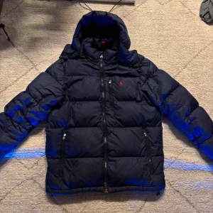 navy blå ralph lauren jacka - Säljer en snygg blå dunjacka från Polo Ralph Lauren i storlek M. Jackan har en dragkedja och är perfekt för kalla vinterdagar. Den är i mycket bra skick och har en klassisk design med en liten röd logga på bröstet. Perfekt för både vardag och fest!
