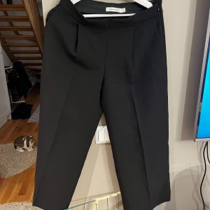 Svarta kostymbyxor från Zara - Säljer ett par stilrena svarta kostymbyxor från Zara. De är normal midja  och har en klassisk rak passform.  Byxan är 3/4 längd . Perfekta för både kontoret och festligare tillfällen. Byxorna är i mycket bra skick och har diskreta fickor baktill.