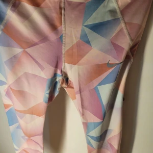 Flerfärgade leggings från Nike - Säljer ett par snygga flerfärgade leggings från Nike i storlek M. De har ett geometriskt mönster i rosa, blått och orange. Perfekta för träning med Dri-FIT material som håller dig torr och bekväm. Midjan är elastisk med Nike-logga för extra stil. Passar perfekt för gymmet eller löprundan!