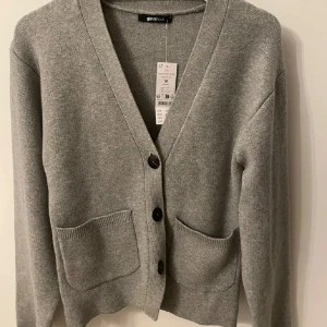 Grå stickad kofta från Gina Tricot - Säljer en superfin grå stickad kofta från Gina Tricot i storlek M. Den har en v-ringad design med knappar framtill och två praktiska fickor. Perfekt för både höst och vinter, och den är i helt nytt skick med prislappen kvar. Passar perfekt för en mysig och stilren look! 🧶