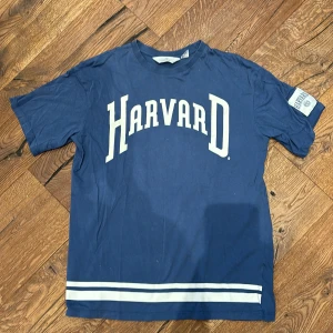 Blå Harvard t-shirt från H&M - Säljer en blå t-shirt från H&M med Harvard-tryck i vitt på framsidan. Den har korta ärmar och två vita ränder längst ner. Perfekt för en avslappnad stil eller för att visa lite college-vibbar. Passar bra till jeans eller shorts!
