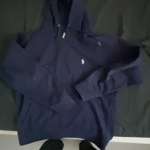 Mörkblå hoodie från Ralph Lauren - Säljer en snygg mörkblå hoodie från Ralph Lauren med dragkedja och den klassiska loggan på bröstet. Perfekt för höst och vinter, med långa ärmar och en skön passform. Perfekt för både vardag och chill , har endast andvänts i 2 månader helt ny i princip från zalando! Pris kan diskuteras