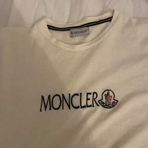 Säljer en snygg vit t-shirt från Moncler i bomull. Den har korta ärmar och Monclers logga broderad på bröstet i svart och rött. Perfekt för en stilren och avslappnad look. Passar både till vardags och fest!