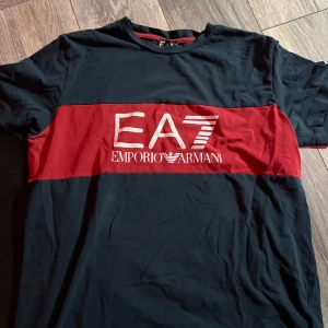 Svart och röd t-shirt från Emporio Armani - Snygg svart t-shirt med röd panel och vit logga från Emporio Armani. Perfekt för en casual look eller att klä upp med en snygg jacka. Den är kortärmad och gjord i ett mjukt material som känns skönt mot huden. Passar både till vardags och fest!
