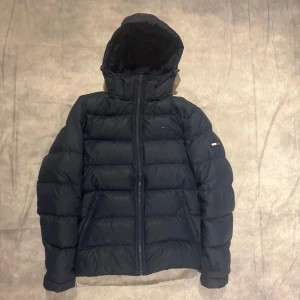 Mörkblå dunjacka från Tommy Hilfiger - Säljer en snygg mörkblå dunjacka från Tommy Hilfiger. Jackan har en dragkedja framtill och en mysig huva. Perfekt för kalla dagar med sin pufferstil och långa ärmar. Den har också det klassiska Hilfiger-emblemet på ärmen. Passar perfekt för både höst och vinter! Storlek M.