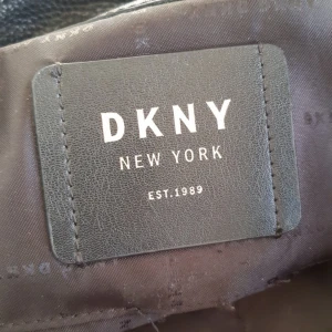 Svart och brun väska från DKNY - Säljer en snygg väska från DKNY i svart och brunt. Den har ett stilrent mönster och är gjord av skinn. Väskan har robusta handtag och små metallfötter på undersidan för extra skydd. Perfekt för både vardag och fest!