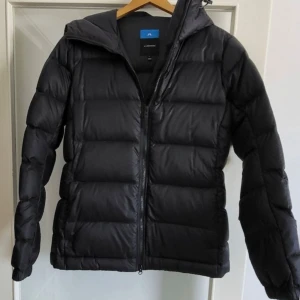 Svart dunjacka från J.Lindeberg - Säljer en svart dunjacka från J.Lindeberg i storlek L. Jackan är i mycket bra skick och perfekt för kalla dagar. Den har en dragkedja framtill och en huva för extra värme. Materialet är 90% dun och 10% fjädrar, vilket gör den både lätt och varm. Perfekt för vinter och höst!