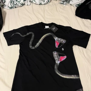 Marcelo Burlon Svart t-shirt med ormmotiv - Säljer en svart t-shirt med ett coolt ormmotiv som slingrar sig över framsidan. T-shirten är kortärmad och har en normal passform. Perfekt för en avslappnad stil eller en utekväll. 🐍