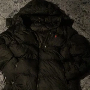 Svart dunjacka från Polo Ralph Lauren - Säljer en snygg svart dunjacka från Polo Ralph Lauren i storlek M. Jackan har en broderad röd logga på bröstet och praktiska dragkedjor. Perfekt för kalla dagar med sin varma och puffiga design. Passar både till vardags och lite finare tillfällen. Inga defekter alls 