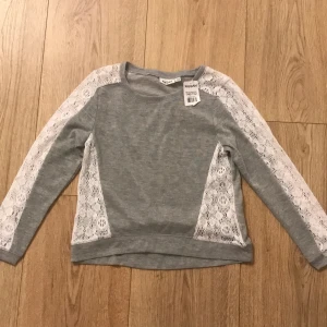 Grå tröja med spetsdetaljer från KappAhl - Säljer en snygg grå tröja från KappAhl med vita spetsdetaljer längs ärmarna och sidorna. Perfekt för en stilren och bekväm look. Tröjan är i nytt skick och passar perfekt för vår och höst. 