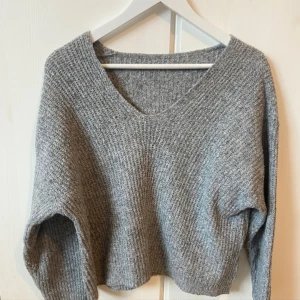 Grå stickad tröja - Mysig grå stickad tröja med en avslappnad passform och v-ringad hals. Perfekt för kyliga dagar och att mysa i hemma. Den har långa ärmar och en croppad stil som ger en modern touch. Passar perfekt till jeans eller leggings!