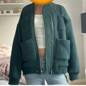 Mörkgrön quiltad bomberjacka - Säljer en snygg mörkgrön quiltad bomberjacka. Den har en dragkedja framtill och stora fickor. Perfekt för höst och vår, med en skön och avslappnad passform. Jackan är långärmad och har ribbade muddar vid ärmslut och nederkant. Storlek XS men oversized!!