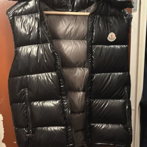 Svart dunväst från Moncler - Säljer en snygg svart dunväst från Moncler i mycket bra skick. Västen har en glansig finish och är perfekt för höst och vinter. Den har en dragkedja framtill och en praktisk huva. Insidan har ett coolt serietidningsmönster. Perfekt för att hålla sig varm och stilren! Är öppen för bud