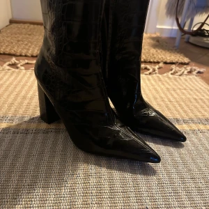 Svarta stövletter med krokodilmönster - Snygga svarta stövletter i skinn med krokodilmönster och spetsig tå. De har en hög klack och dragkedja på sidan för enkel på- och avtagning. Perfekta för en stilren look till både vardag och fest!