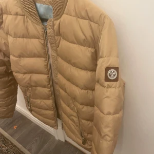 Beige pufferjacka med dragkedja - Säljer en snygg beige pufferjacka från Morris. Jackan har en dragkedja framtill och två fickor med dragkedjor. Den är perfekt för höst vår och vinter, och har en stilren design med ett märke på ärmen. Superbra skick aldrig använd bara provad och riktigt varm!
