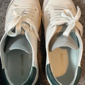 Vita sneakers från Axel Arigato - Säljer ett par snygga vita sneakers från Axel Arigato med gröna detaljer och en cool broderad fågel på sidan. Skorna har snörning och är i mycket bra skick. Perfekta för vardagen, priset går att diskutera 