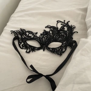 Svart spetsmask - Säljer en elegant svart mask i spets med intrikata detaljer. Perfekt för maskerad eller fest! Den har band för att knyta runt huvudet och ger en mystisk look. Passar perfekt till en kväll med glamour och mystik. (Aldrig använd, köpt för 99kr på partyland)