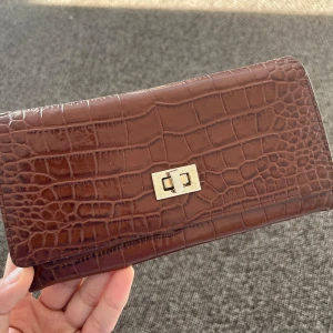 Brun plånbok från GANT - Snygg brun plånbok i krokodilpräglat skinn från GANT. Den har en elegant låsdetalj i guld och flera fack för kort och kontanter. Perfekt för att hålla ordning på dina värdesaker med stil.
