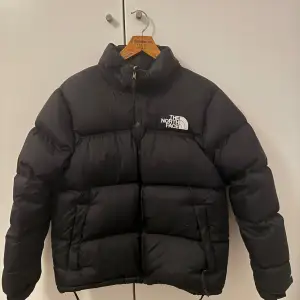 Säljer en svart pufferjacka från The North Face i mycket bra skick. Jackan är perfekt för kalla vinterdagar med sin tjocka vaddering och långa ärmar. Den har en dragkedja framtill och en hög krage för extra värme. Loggan syns tydligt på bröstet och ryggen. Passar både till vardags och utomhusaktiviteter.