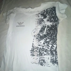 Vit t-shirt från Emporio Armani - Säljer en vit t-shirt från Emporio Armani med ett coolt svart grafiskt tryck på ena sidan. T-shirten har korta ärmar och en klassisk rund halsringning. Det finns en liten fläck vid slutet av framsidan, men annars i bra skick. Perfekt för en avslappnad stil! 