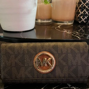 Brun plånbok från Michael Kors - Snygg brun plånbok från Michael Kors med deras ikoniska logotyp i metall på framsidan. Plånboken har ett mönster av MK-loggor och stängs med en tryckknapp. Insidan har flera fack för kort och en dragkedja för mynt. Perfekt för att hålla ordning på dina saker med stil!