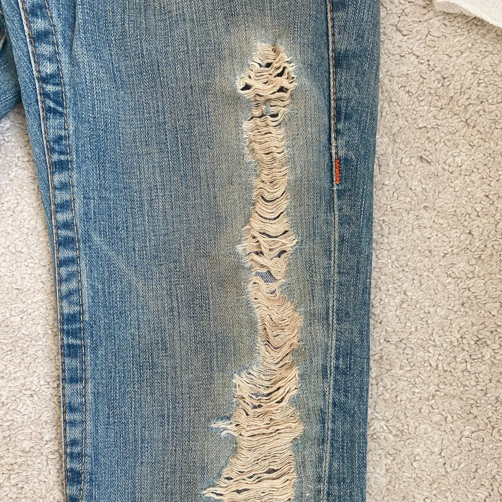 Säljer ett par snygga blå jeans från True Religion i storlek 27. De har en cool slitning på framsidan och en bootcut-stil. Midjemåttet är 39 cm. Har en liten fläck (bild 3). Lite slitna vid hälarna.❤️Kolla min profil för andra jeans.😍😍😍😍. Jeans & Byxor.