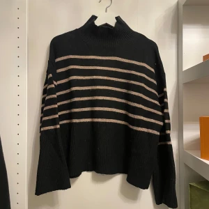 Svart och beige randig stickad tröja - Säljer en snygg svart stickad tröja med beige ränder. Den har en hög krage och långa ärmar, perfekt för kyliga dagar. Tröjan är i mycket bra skick och passar både till vardags och lite finare tillfällen. Supermysig och stilren!