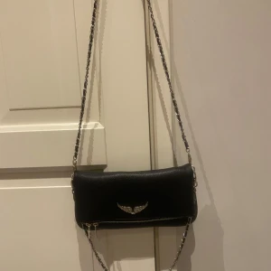 Svart axelväska från Zadig & Voltaire - Snygg svart axelväska i skinn från Zadig & Voltaire. Väskan har en kedjerem och ett coolt emblem på framsidan. Perfekt för både vardag och fest! Den har en dragkedja för säker förvaring. Ordinarie pris ca 4000kr