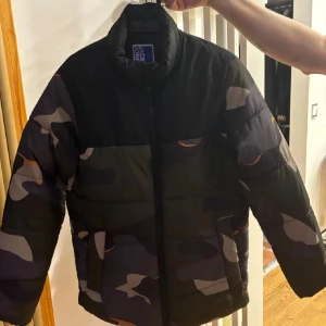 Camouflage pufferjacka från Jack & Jones - Säljer en cool pufferjacka från Jack & Jones i ett snyggt camouflagemönster. Jackan är perfekt för höst och vinter med sin quiltade design och dragkedja framtill. Den har långa ärmar och är i mycket bra skick. Perfekt för att hålla sig varm och stilren! 🧥