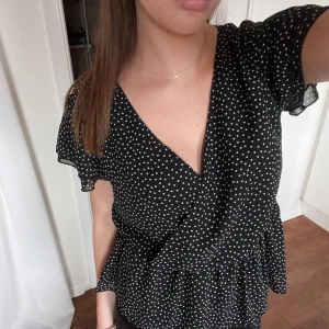 Svart prickig omlottblus - Säljer en superfin svart omlottblus med vita prickar. Blusen har korta ärmar och en smickrande v-ringning. Perfekt för både vardag och fest! 🖤