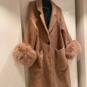 Brun kappa med päls från Meotine - Säljer en superfin brun kappa med stora, fluffiga äkta pälsdetaljer vid ärmsluten. Den har en klassisk design med knappar framtill och stora fickor. Perfekt för höst och vinter när du vill hålla dig varm och stilren. Passar både till vardags och festliga tillfällen. 🧥✨en knapp saknas på ena ärmen men syns inte när man har på sig den!