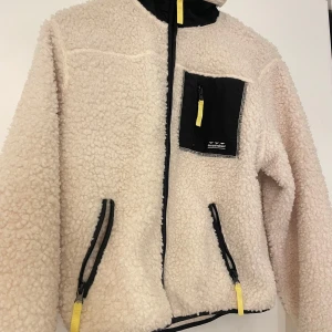 Beige teddyjacka med dragkedja - Mysig beige teddyjacka med svart och gul detalj. Den har en praktisk dragkedja framtill och en bröstficka med dragkedja. Perfekt för kyliga dagar och ger en cool look med sina kontrasterande detaljer.