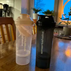 Två praktiska Smartshake shakers i svart och rosa. Perfekta för träning och att blanda proteinshakes. De har en smart design med skruvlock och inbyggd sil för att undvika klumpar. Lätta att rengöra och ta med sig till gymmet eller på språng.