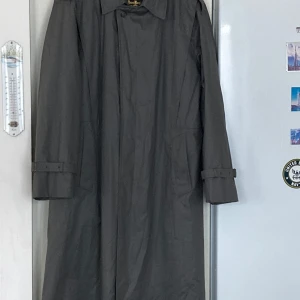 Grå trenchcoat från Dress Bocaj of Sweden  - Säljer en elegant grå trenchcoat från Dress Bocaj of Sweden. Den har en klassisk design med bälte i midjan och långa ärmar. Perfekt för höst och vår. Jackan är i mycket bra skick och gjord av bomull, vilket gör den både bekväm och stilren.