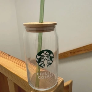 Starbucks glasburk med sugrör - Snygg och praktisk glasburk från Starbucks med ett grönt sugrör och trälock. Perfekt för kalla drycker som iskaffe eller iste. Den har Starbucks-loggan tryckt på framsidan och texten 'Chilled Coffee'. Perfekt för att ta med sig på språng eller njuta av hemma.