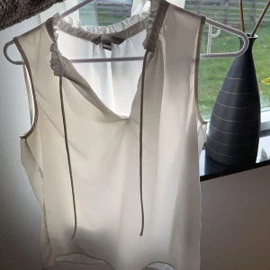 Vit ärmlös blus - Säljer en stilren vit ärmlös blus med knytband vid halsen. Perfekt för både vardag och fest. Blusen har en luftig passform och är gjord i ett lätt material som passar bra till vår och sommar.