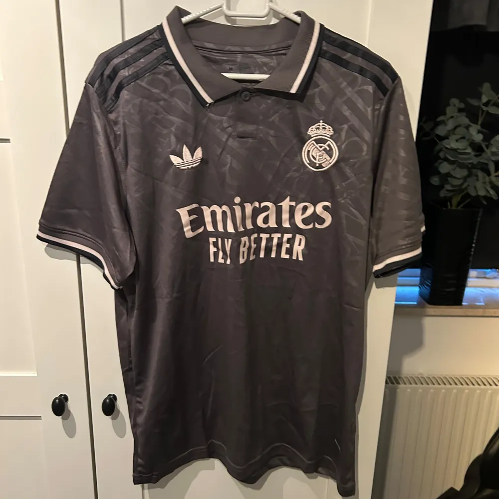 Säljer en snygg grå Real Madrid tröja från Adidas i nytt skick. Den har korta ärmar och ett stilrent mönster. Perfekt för alla fotbollsfans! 🖤. T-shirts.