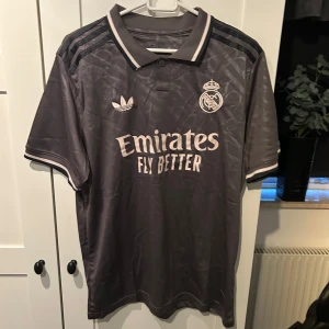 Grå Real Madrid fotbollströja från Adidas - Säljer en snygg grå Real Madrid tröja från Adidas i nytt skick. Den har korta ärmar och ett stilrent mönster. Perfekt för alla fotbollsfans! 🖤