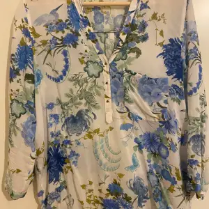 Blus med blommigt mönster i blått och grönt på vit bakgrund. Blusen har en v-ringning och knappar framtill. Den har även en ficka på bröstet. Ärmarna är tre fjärdedels långa.