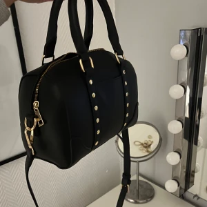 Svart axelväska/handväska med gulddetaljer - Snygg svart axelväska från Even&Odd med gulddetaljer och dragkedja. Väskan har både handtag och en justerbar axelrem så man kan bära den olika. Väldigt rymlig och som i nyskick💕