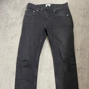 Svarta jeansbyxor från 157 - Svarta jeansbyxor från 157 med klassisk design. De har en dragkedja och knapp framtill samt fem fickor. Perfekta för en stilren look.                   Storlek: W:28 L:32                                                                 -                                   (OBS! Byxorna har vikningsveck med viss färgskillnad som syns om de inte viks upp. Påverkar ej kvalitet eller passform. Se sista bild.)