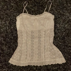 Vit spetslinne - Säljer en söt vit spetslinne med tunna axelband och vacker spetsdetalj. Linnet har ett blommigt mönster och en lätt volangkant nedtill. Perfekt för varma dagar eller som ett lager under en tröja.