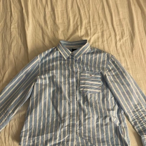 Blå och vit randig skjorta från GANT - Säljer en stilren blå och vit randig skjorta från GANT. Skjortan har långa ärmar och en bröstficka med broderad logotyp. Perfekt för en klassisk look.