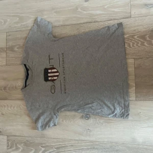 Grå t-shirt från GANT - Säljer en grå t-shirt från GANT med tryck på framsidan. T-shirten har korta ärmar och är tillverkad i ett mjukt material. Perfekt för en avslappnad stil.